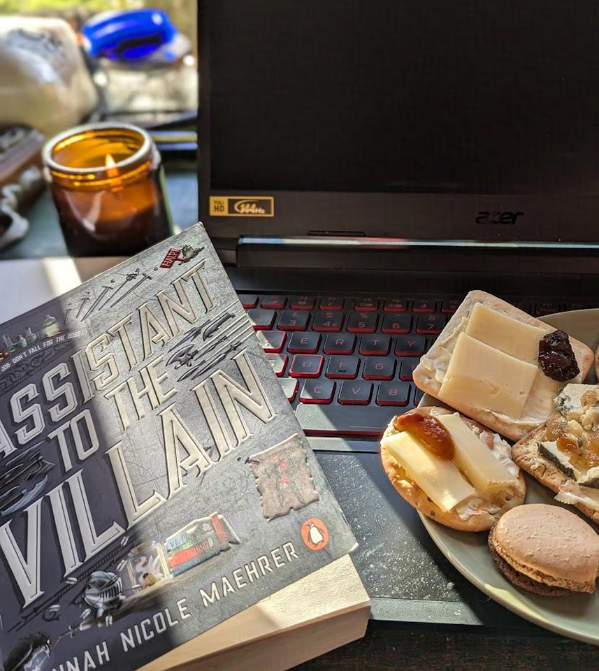 My laptop sat on my desk, with a plate of cheese and crackers. There's a candle in the background, and the book 'Assistant to the Villain' by Hannah Nicole Maehrer. 
