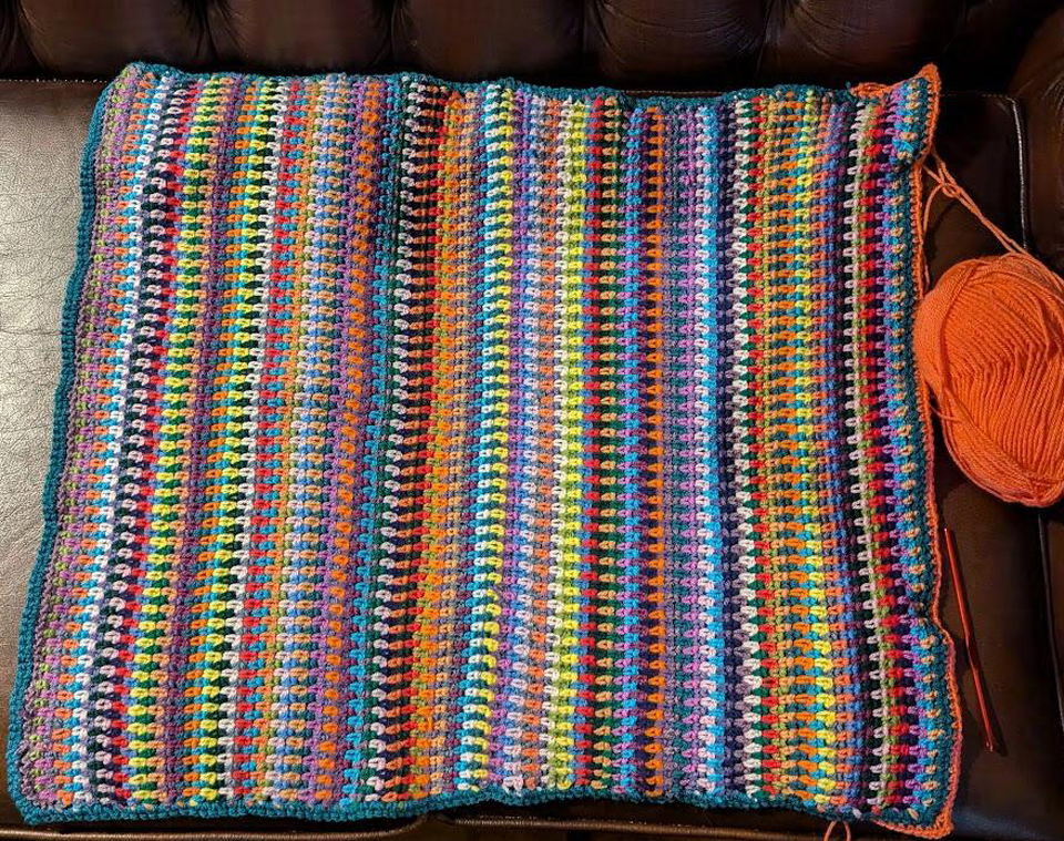 A rectangular crocheted baby blanket made of striped of different colour wool. It's not quite finished here, the edging has just been started. 