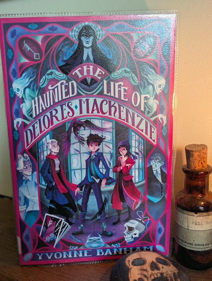 The cover of the book 'The Haunted Life of Delores Mackenzie'. The cover is shades of pink and purple against blue, black and white. The book is standing on a wooden mantelpiece next to an old glass bottle, brown with a cork, and a wooden pebble with a pyrography skull burnt onto it. 