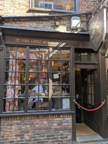 The exterior of The York Ghost Merchant, a very small shop in the quaint, cobbled street called the Shambles. The shopfront is brick, with black paintwork. A red rope is across the door, the shop is so small only a few people are allowed in at a time. 
